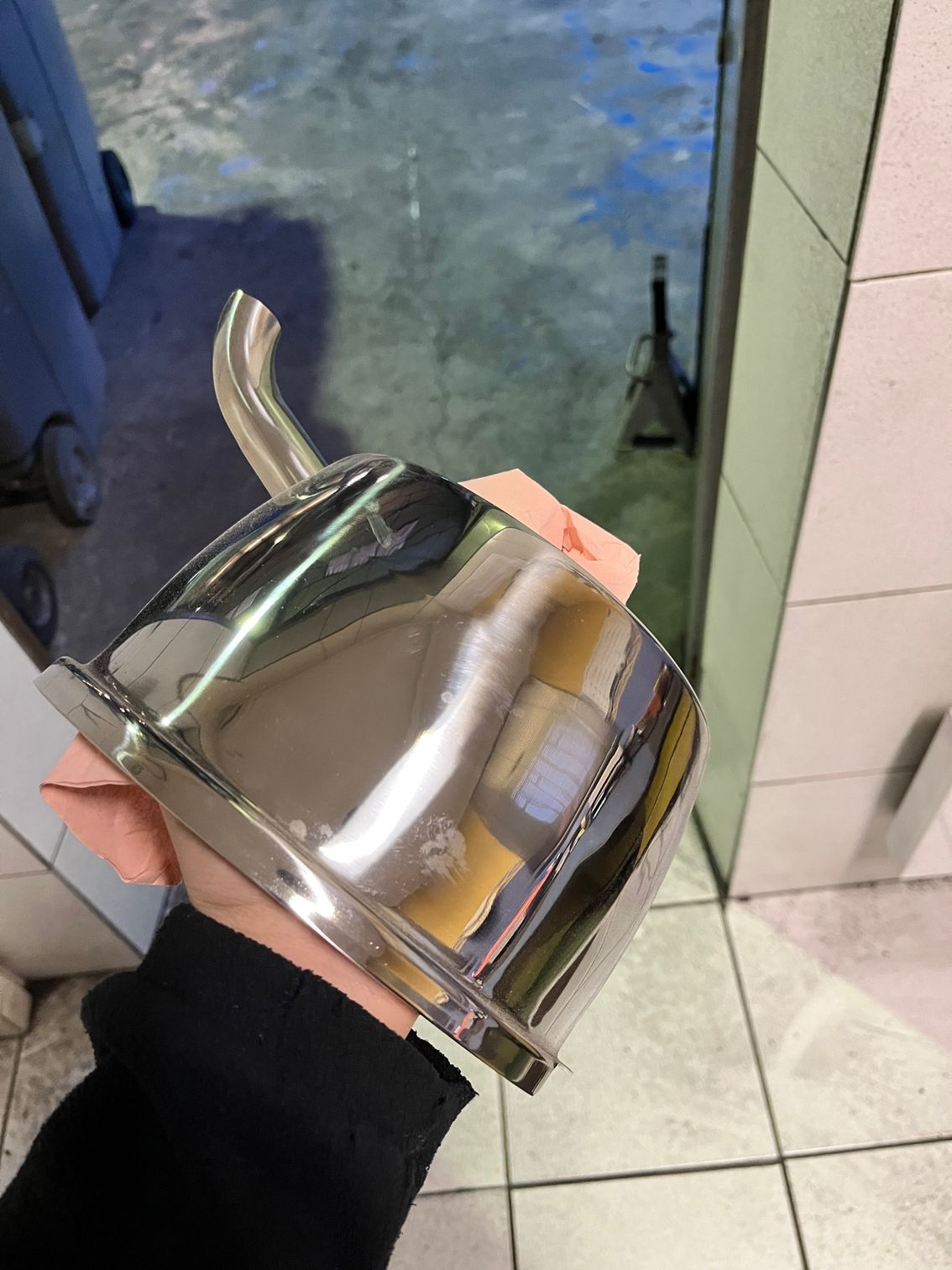 Une main tenant un entonnoir en acier inoxydable brillant, muni d'un bec coudé, dans une pièce au sol carrelé.