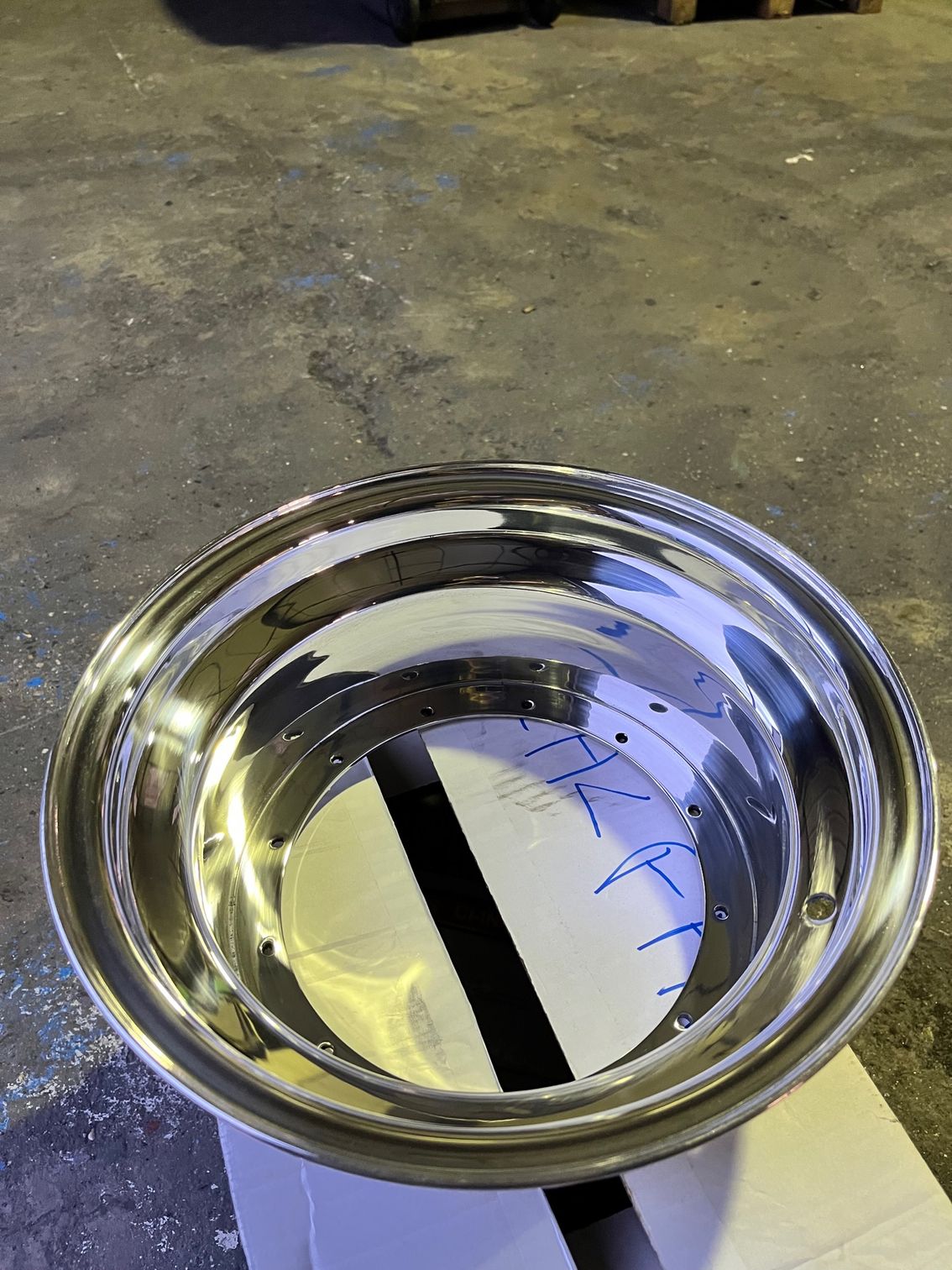 Jante de roue en métal poli brillant posée sur un sol en béton. La jante est circulaire, composée de plusieurs couches et d'un insert en papier.