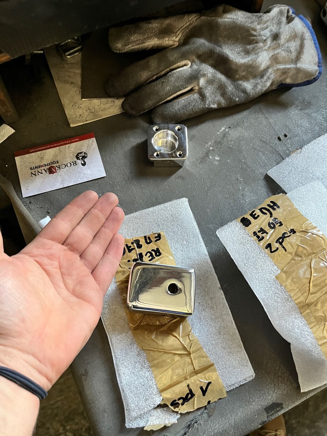 Une main désigne une pièce métallique usinée. D'autres pièces, des gants et des étiquettes sont posés sur un établi.