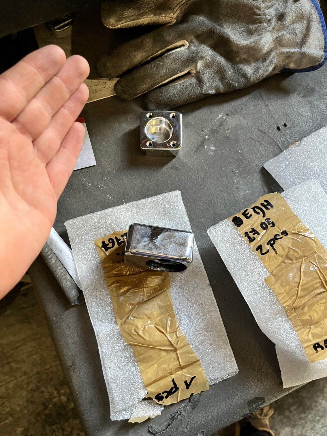 Une main désigne des cubes de métal, l'un d'eux posé sur une table avec des gants et des emballages.