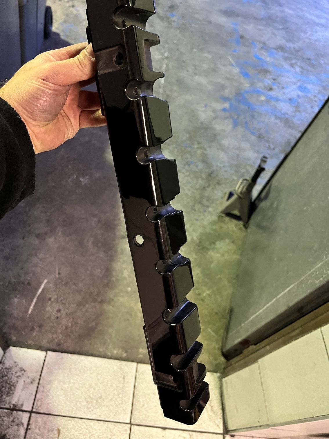 Main tenant une pièce métallique noire brillante présentant des protubérances rectangulaires régulièrement espacées et un trou de fixation.