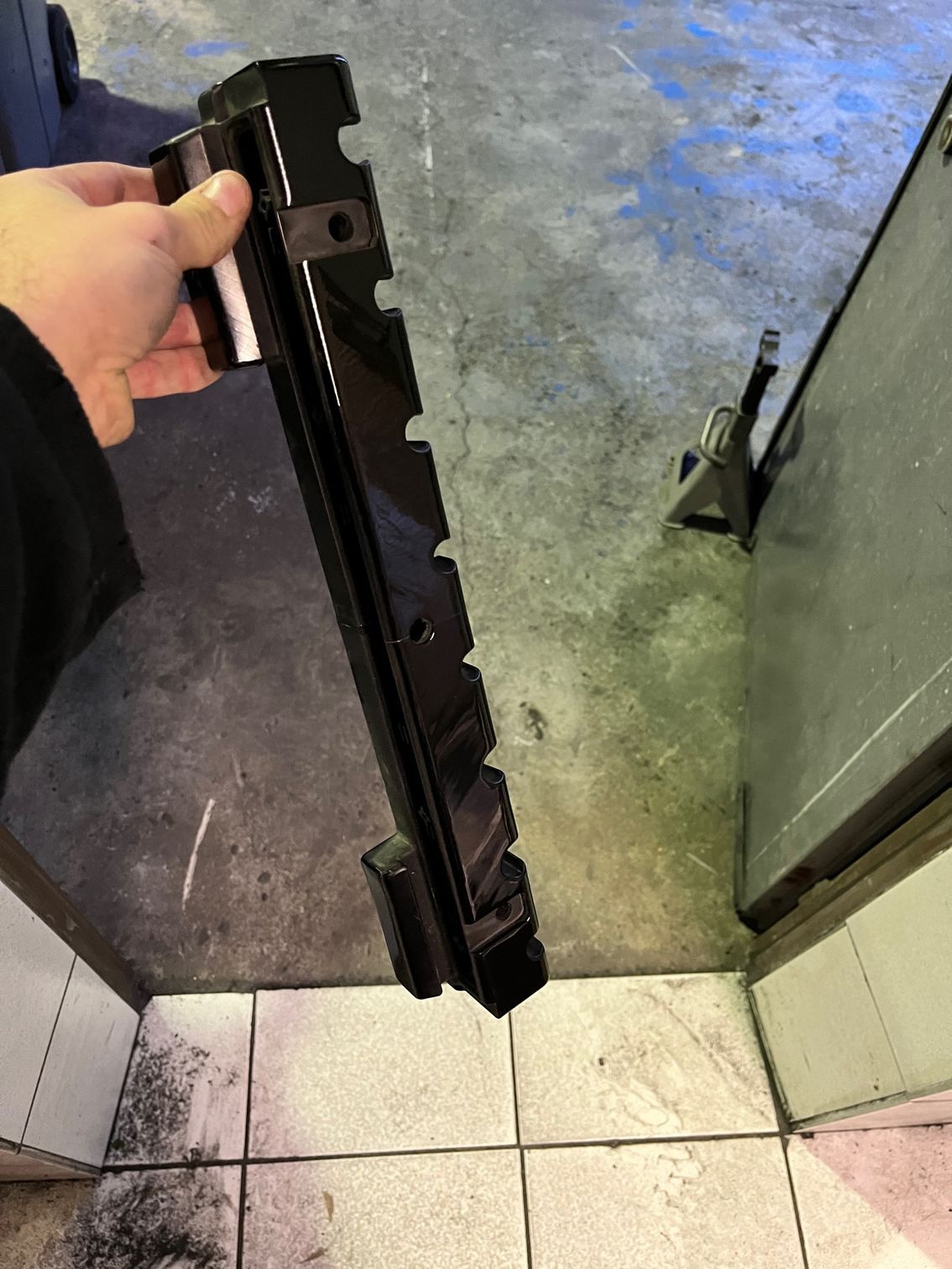 Main tenant une longue pièce métallique noire pour véhicule, munie d'un support de fixation, dans un atelier.