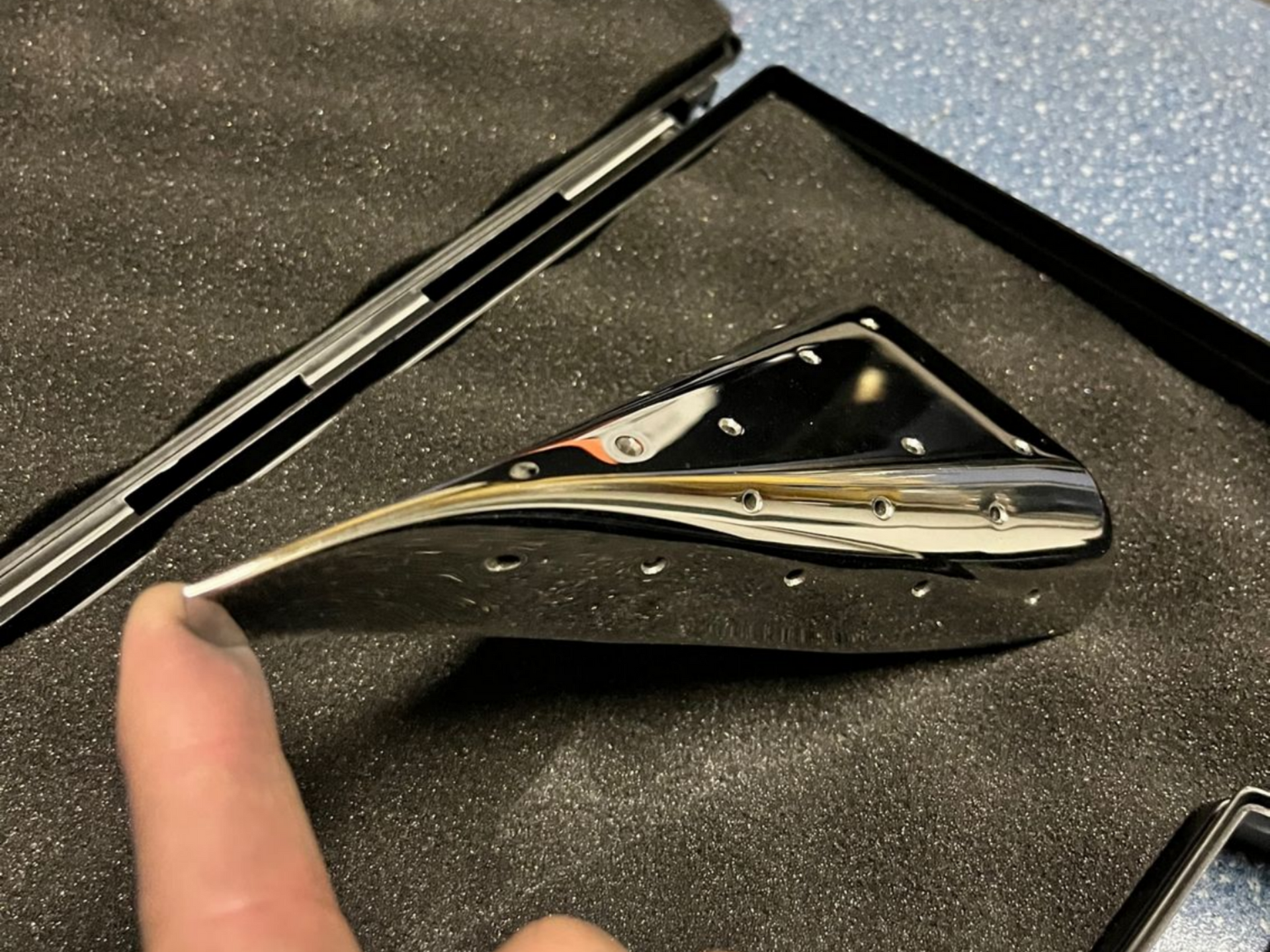 Panneau métallique ovale et brillant percé d'un trou rond. La lumière réfléchie crée des motifs rayés et des reflets bleus.