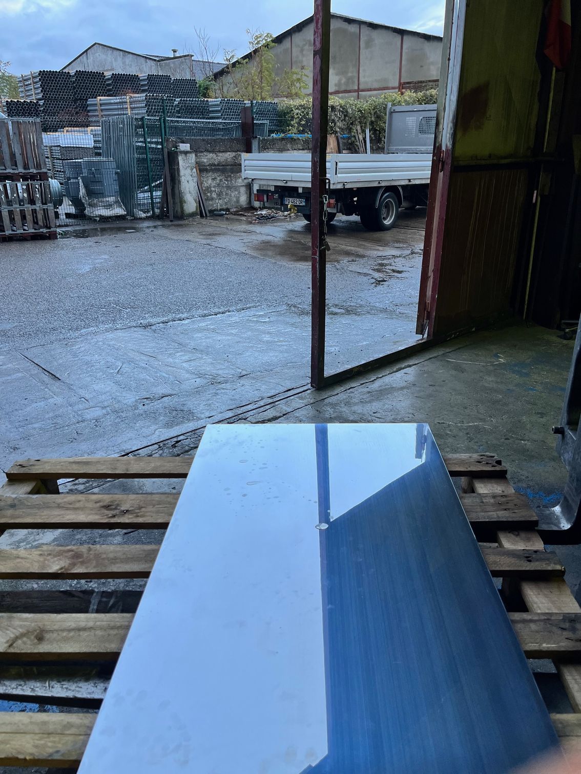 Une plaque de métal brillante et réfléchissante repose sur une palette en bois dans une zone industrielle où l'on aperçoit un camion et des bâtiments.