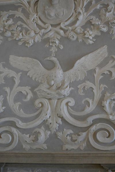 Scultura a rilievo bianca raffigurante un uccello con le ali spiegate, immersa in un elaborato motivo floreale su una parete grigia.