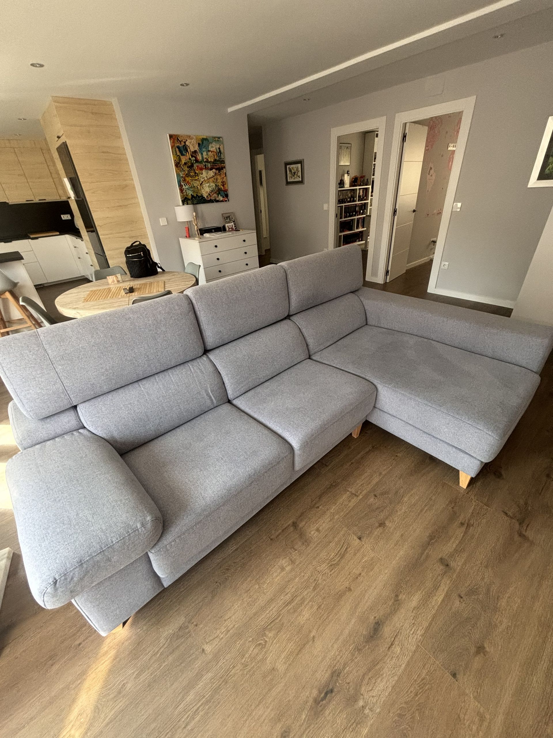 Sofá modular gris en una luminosa sala de estar con suelo de madera y una cocina al fondo.