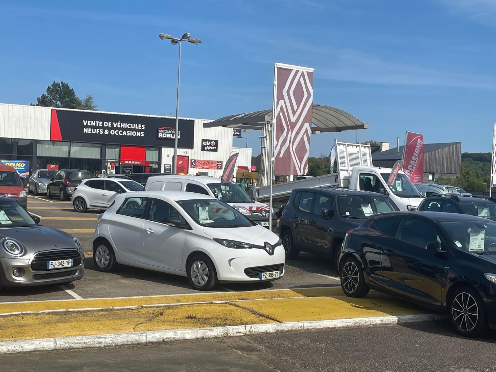 Extérieur du concessionnaire Renault avec des voitures exposées ; voitures bleues, rouges et blanches garées à l'extérieur du bâtiment sous un ciel nuageux.