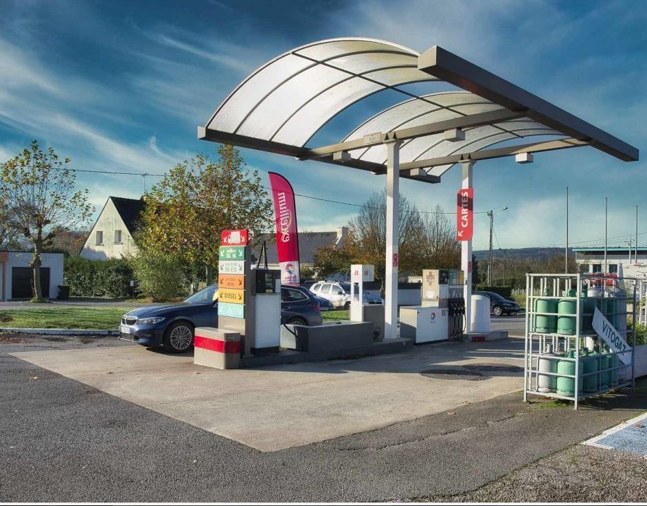 Station-service avec une voiture à une pompe sous un auvent, avec des panneaux et un ciel bleu.