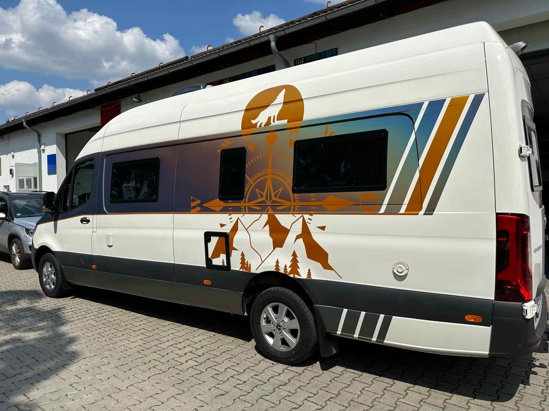 Ein weißes Wohnmobil mit braunen Aufklebern von Bergen, Bäumen und einem Wolf auf einem Kompass.