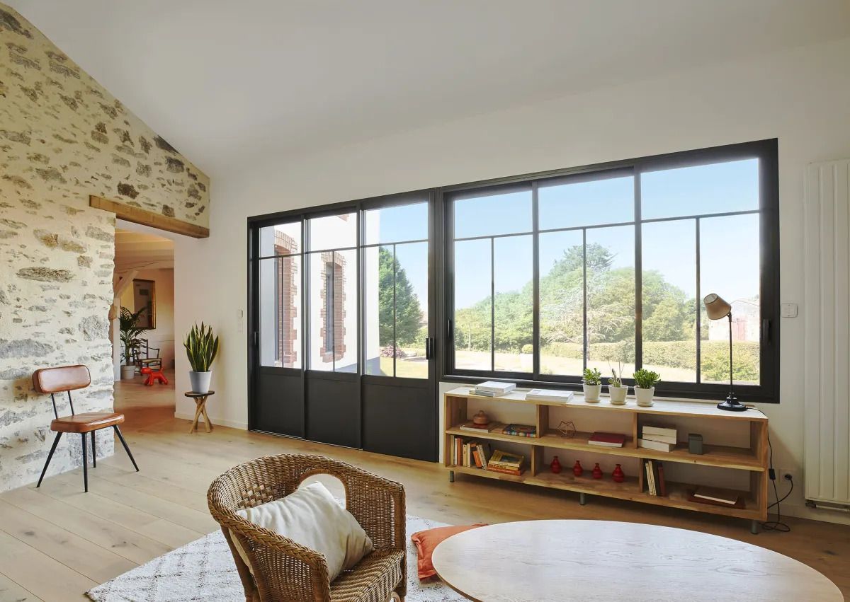 Intérieur d'un salon avec de grandes fenêtres à cadre noir donnant sur une cour verte et un mur de pierre.