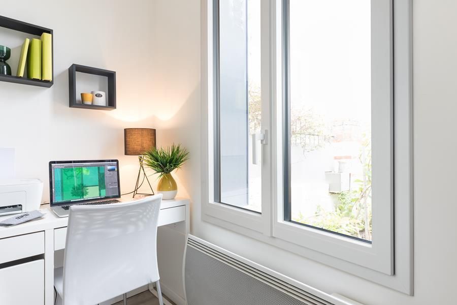 Espace de travail avec bureau, ordinateur portable, fenêtre, lampe et étagères. Murs blancs, lumière naturelle éclatante.