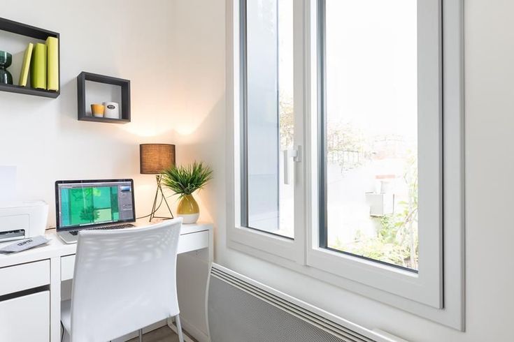 Espace de travail avec bureau, ordinateur portable, fenêtre, lampe et étagères. Murs blancs, lumière naturelle éclatante.