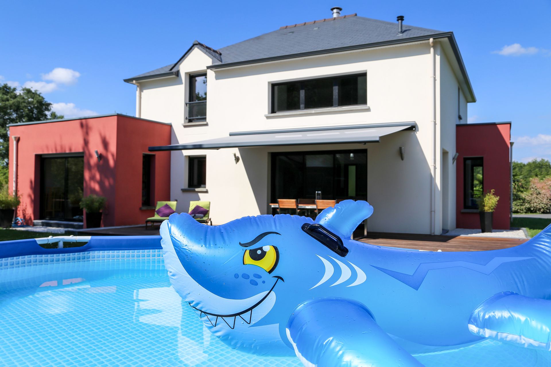 Jardin avec piscine et maison. Un requin gonflable flotte dans la piscine. Maison rouge et blanche sous un ciel bleu.