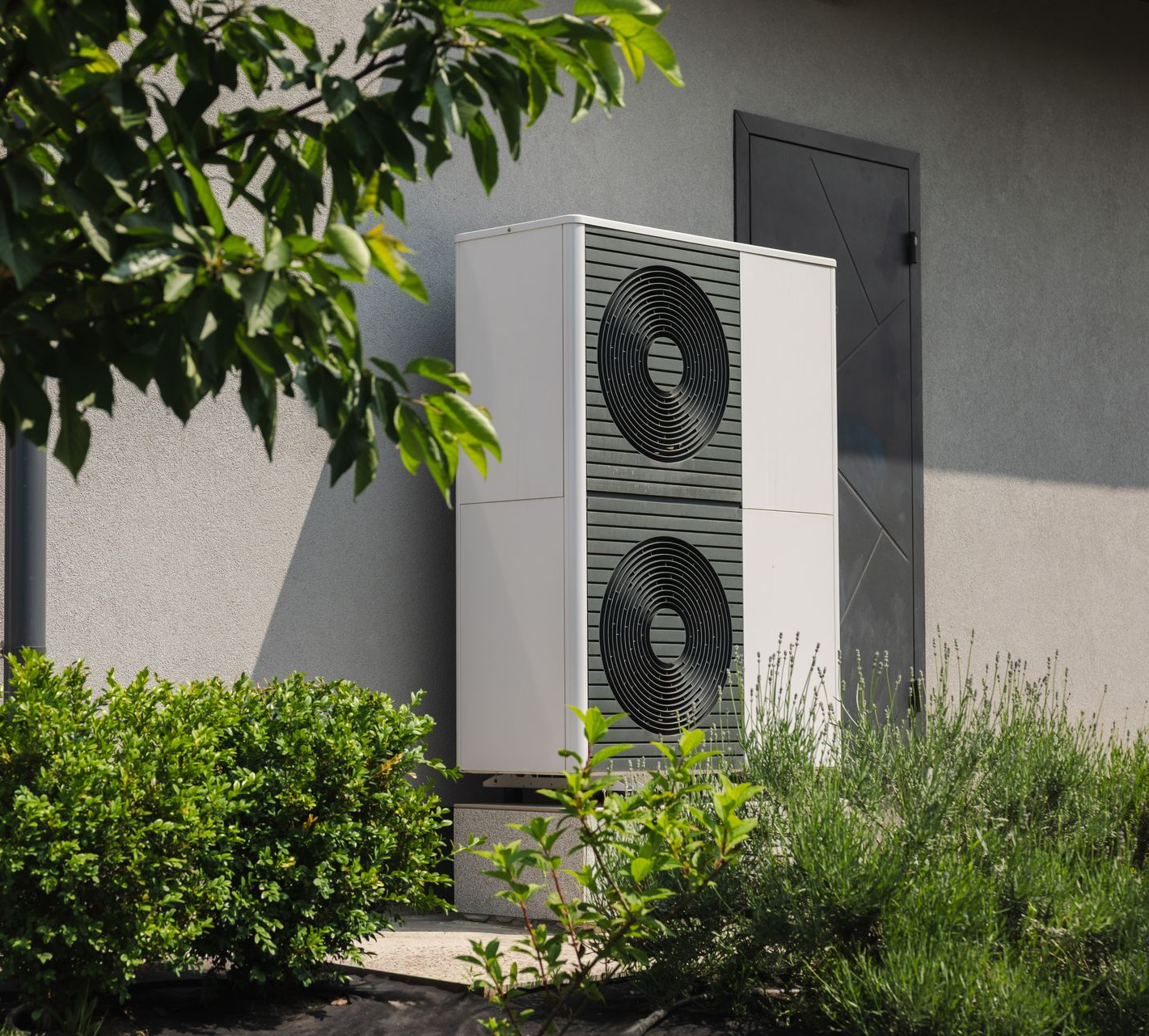 Unité de pompe à chaleur à source d'air montée à l'extérieur d'un bâtiment, partiellement masquée par la verdure.