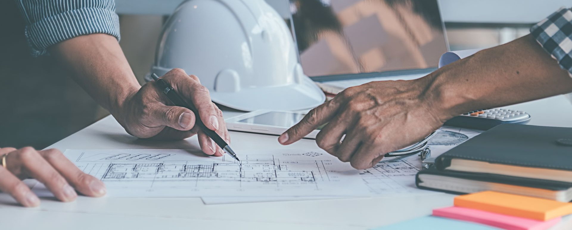 Deux personnes discutent de plans d'architecture à une table. L'une pointe du doigt tandis que l'autre tient un stylo.