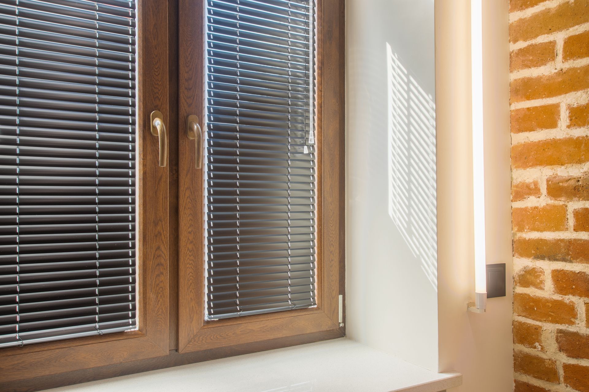 Fenêtre avec stores foncés fermés, cadre marron et mur en briques. La lumière du soleil illumine le mur.