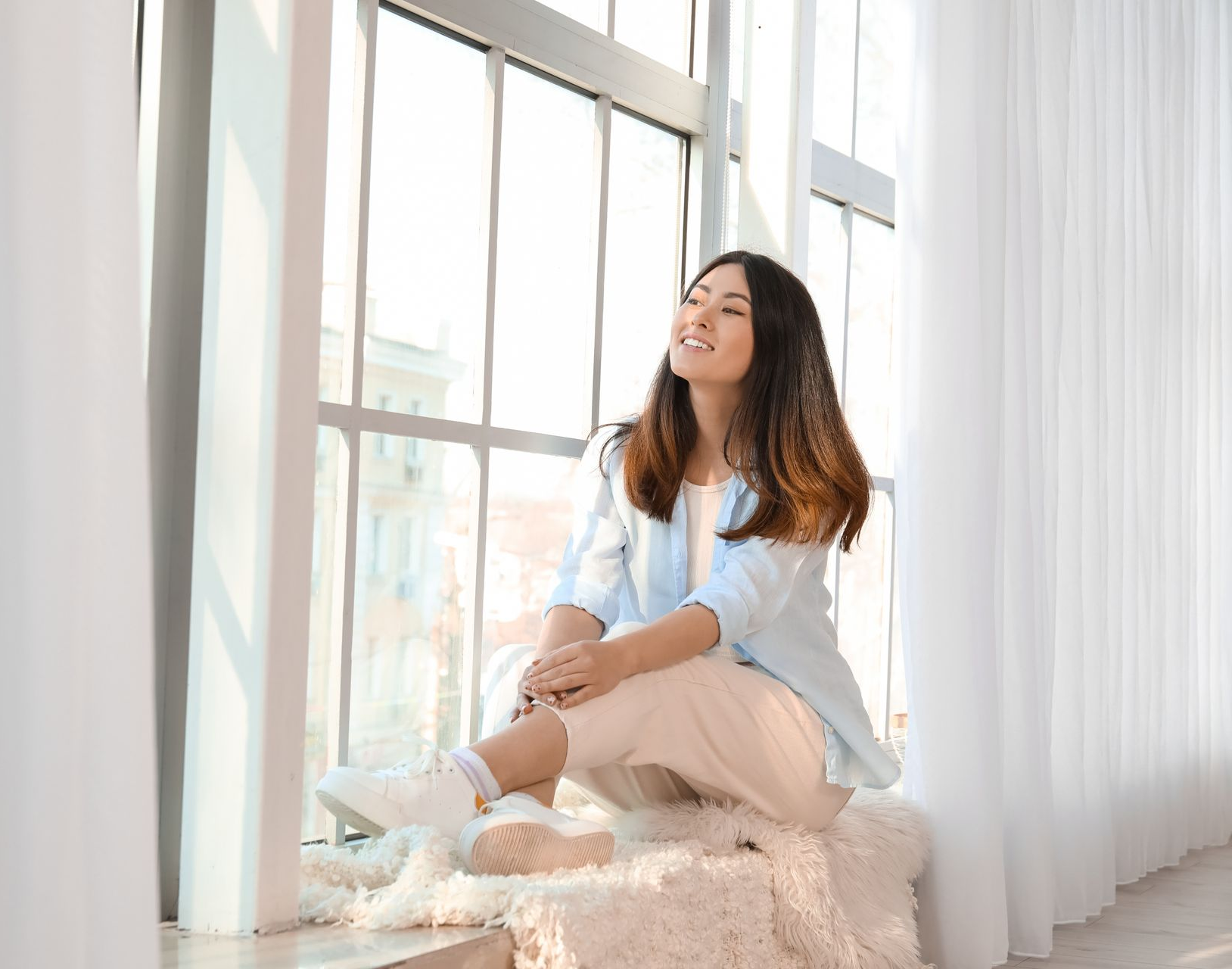 Femme assise près de la fenêtre, souriante ; vêtements de couleur claire, soleil.