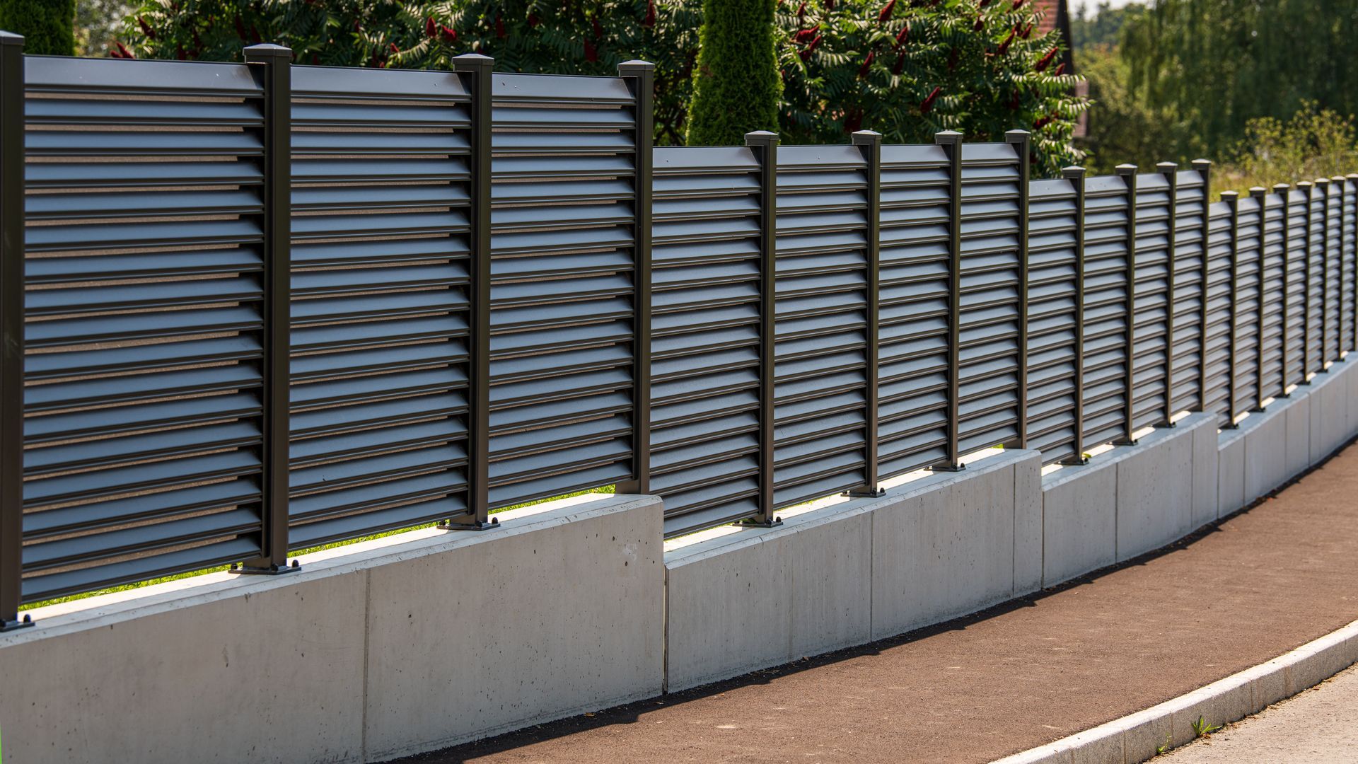 Clôture à lattes gris foncé sur une base en béton, bordant une route.