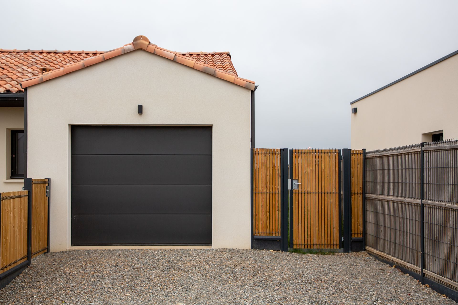 Garage avec porte grise fermée, extérieur beige et toit en tuiles orange, allée en gravier, clôture en bambou et bois.