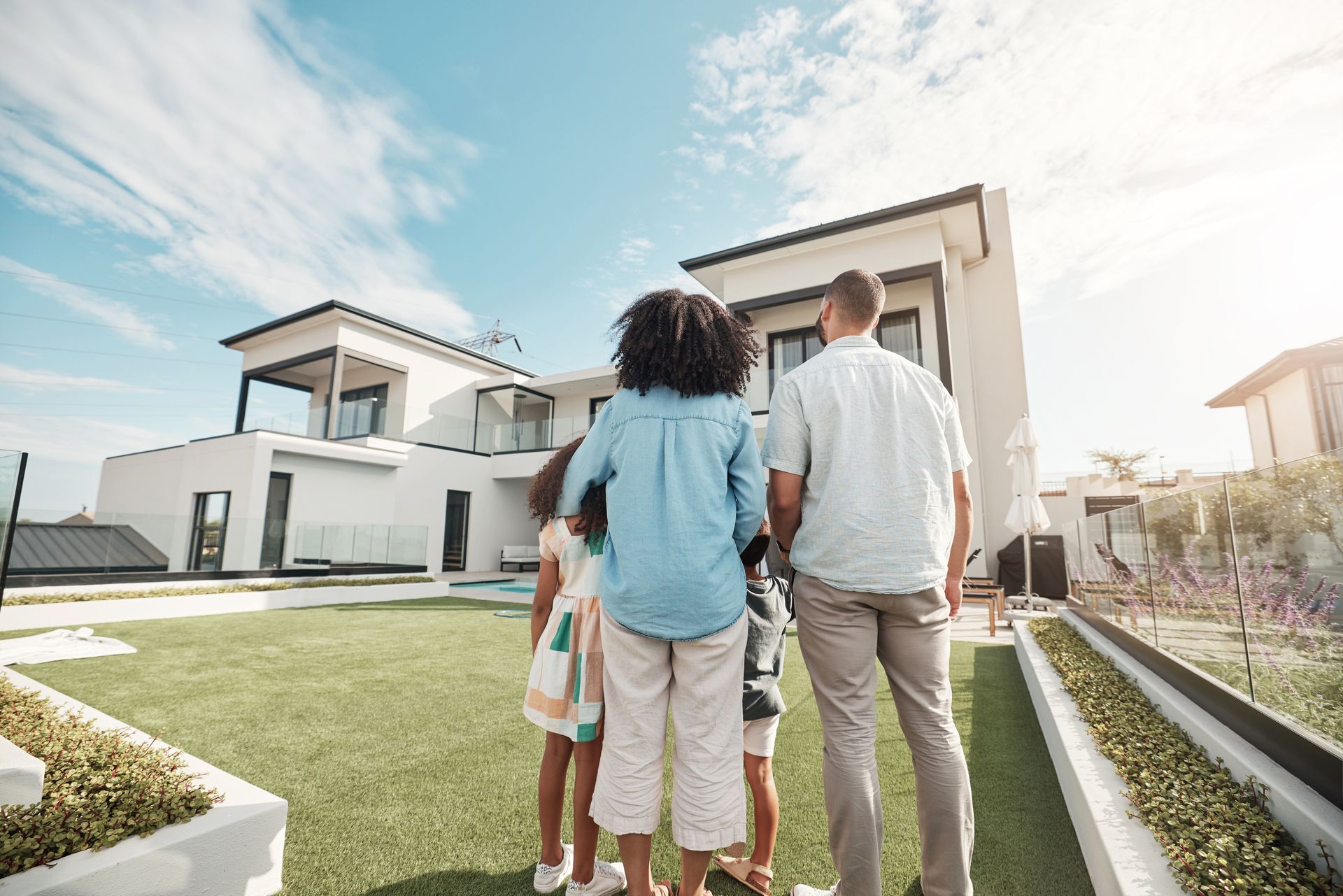 Famille regardant une maison blanche moderne avec une pelouse verte sous un ciel ensoleillé.
