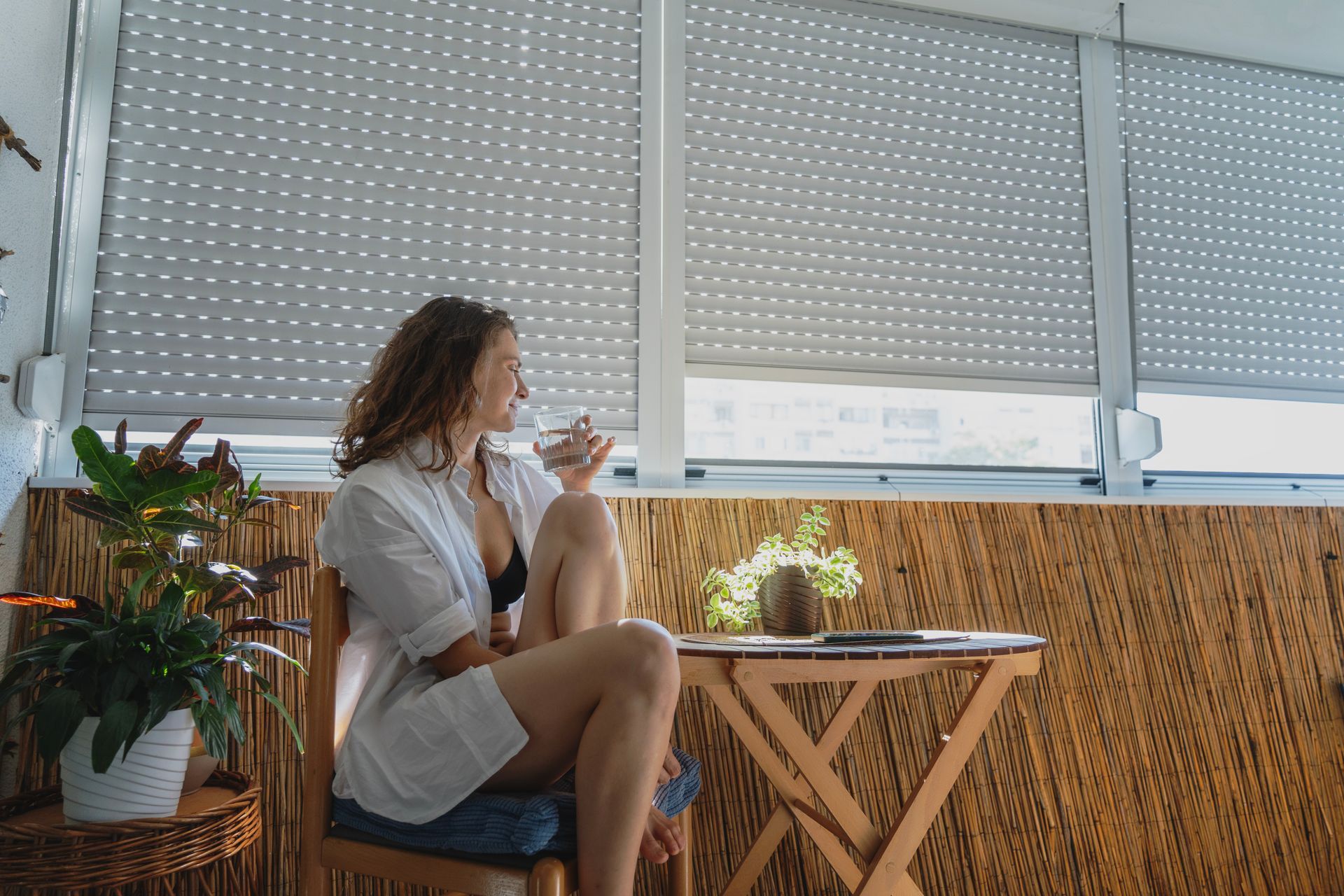Une femme sur un balcon sirote un verre. Vêtue d'un peignoir, assise près d'une petite table et de plantes.