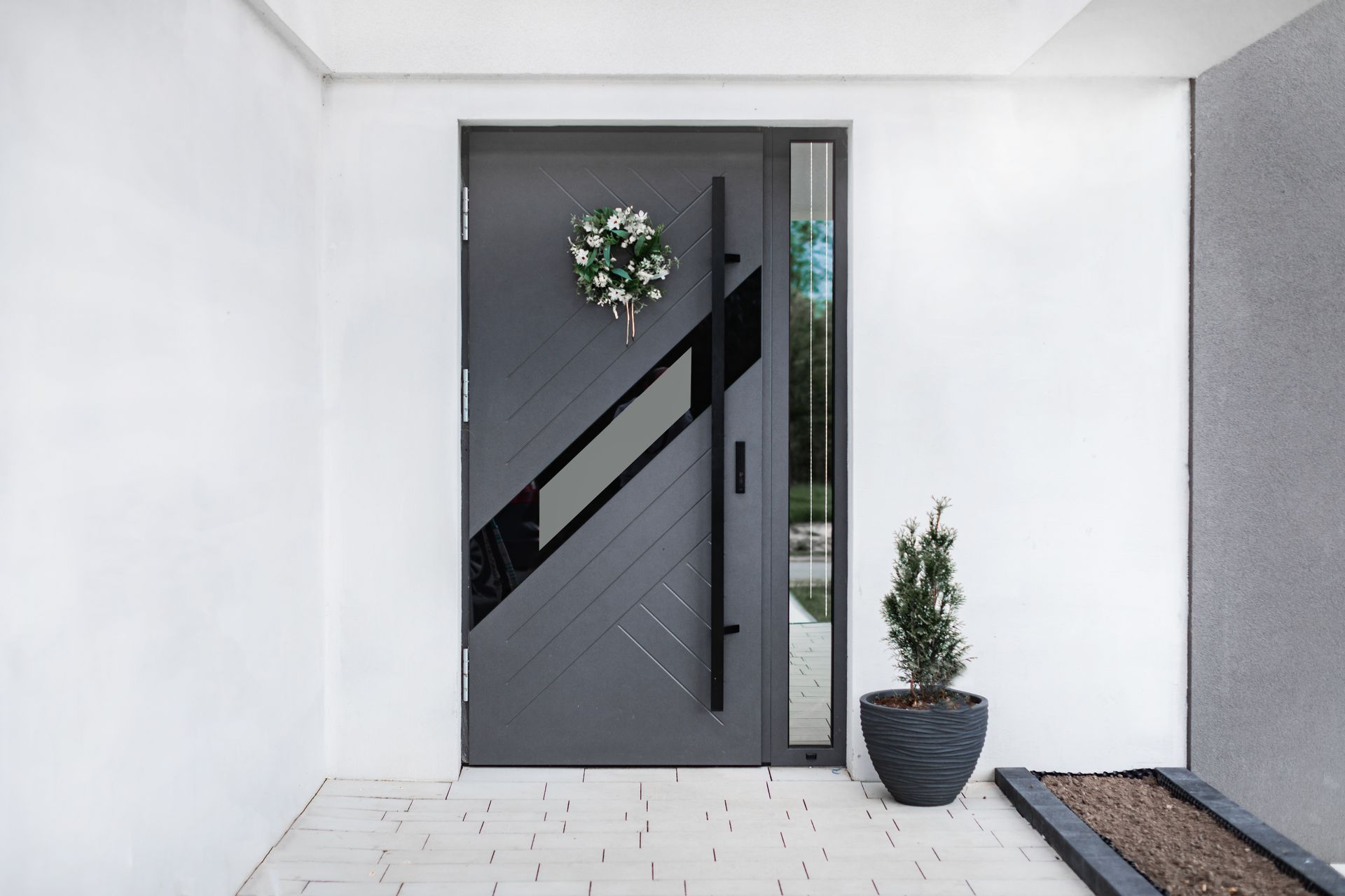 Porte d'entrée grise avec motif diagonal noir, couronne, poignée haute et plante en pot.