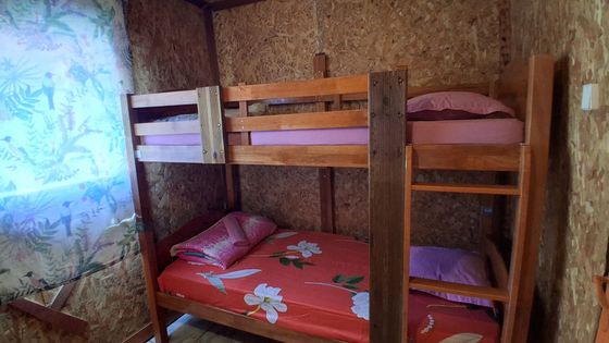 Lit superposé en bois dans une petite chambre avec linge de lit à fleurs et une fenêtre avec un voilage.