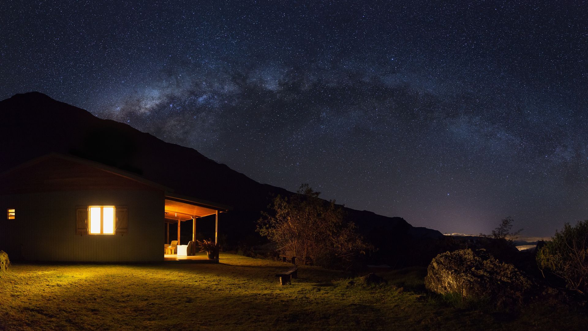 Une cabane éclairée sous un ciel nocturne étoilé, à côté de montagnes sombres et d'un champ.