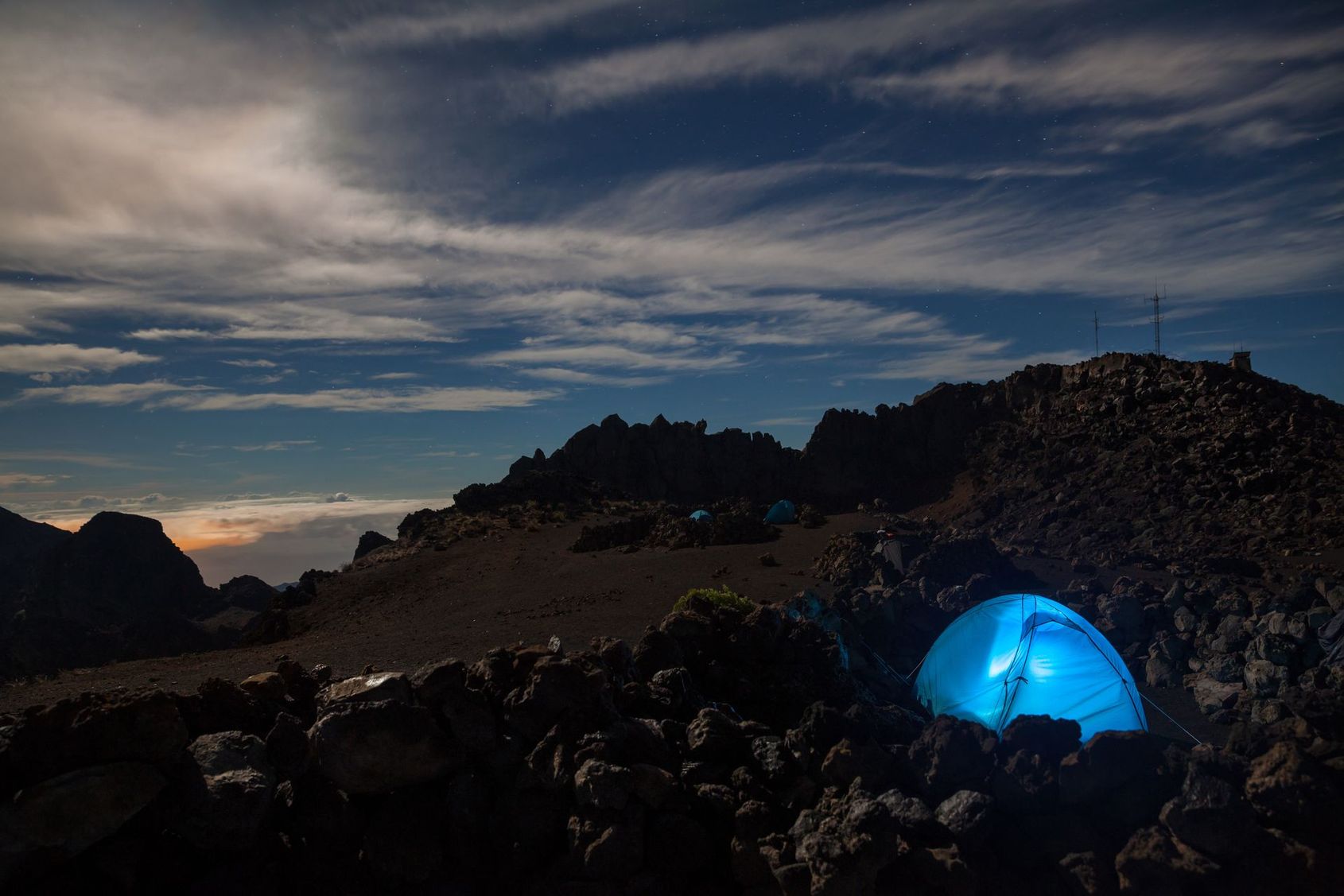 Une tente bleue illuminée sur une colline rocheuse au crépuscule, sous un ciel spectaculaire.