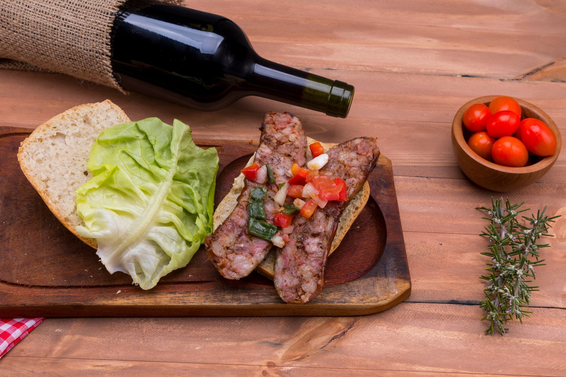 Du vin, une saucisse coupée et de la laitue sur une planche en bois avec des tomates cerises et du romarin.