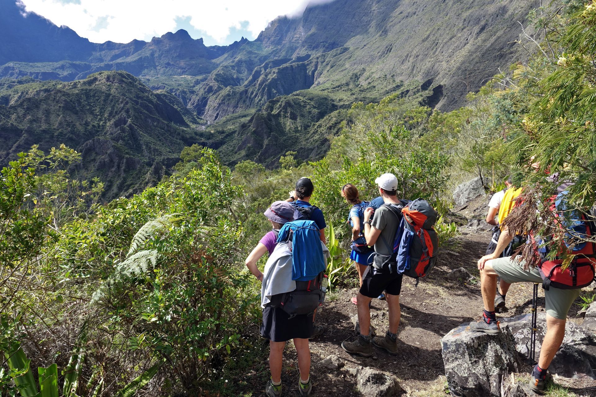 Des randonneurs avec des sacs à dos contemplent une vallée montagneuse luxuriante depuis un sentier rocailleux.