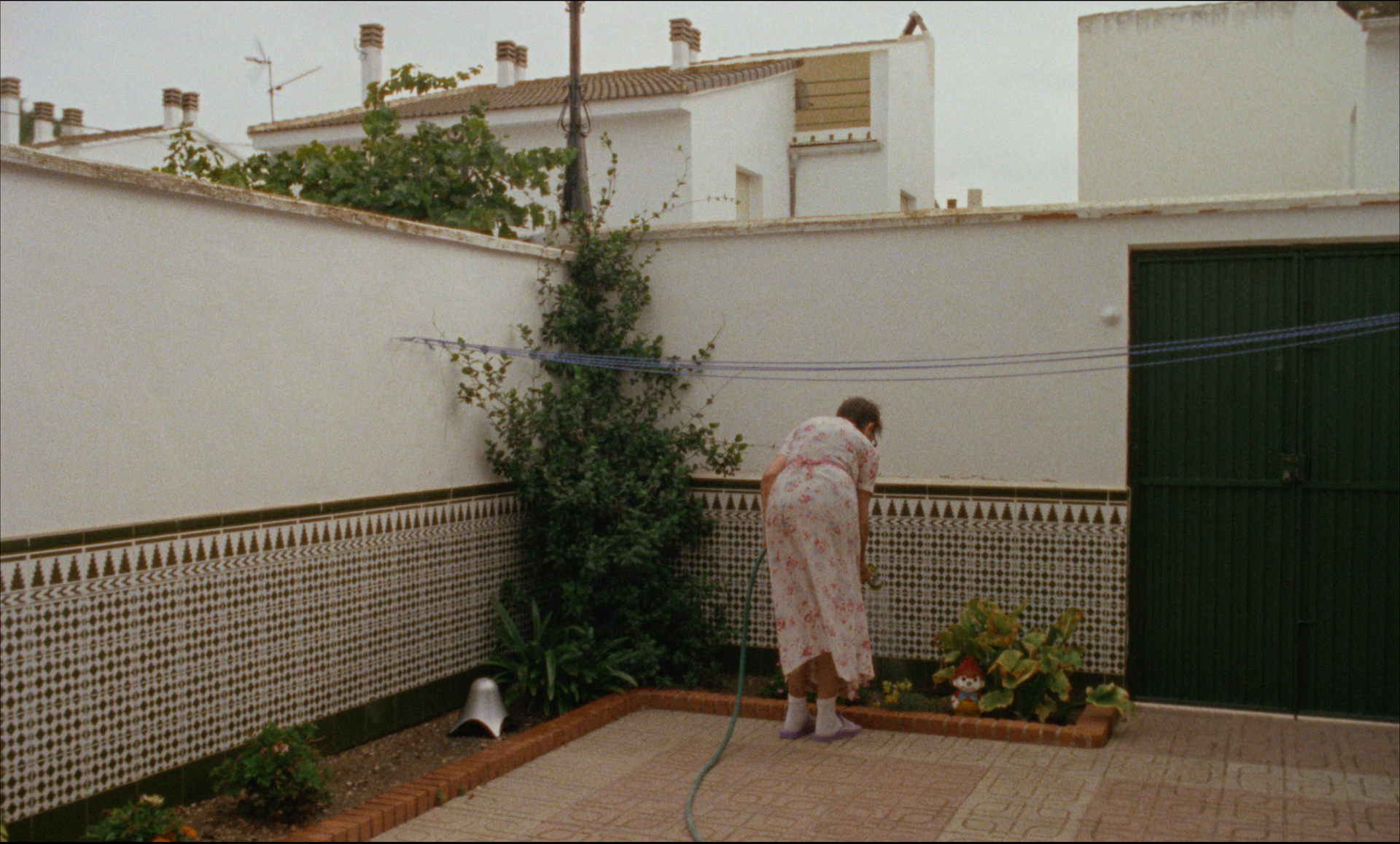 Mujer vestida cuidando plantas en un patio trasero cercado con puerta verde, paredes blancas y detalles de azulejos.