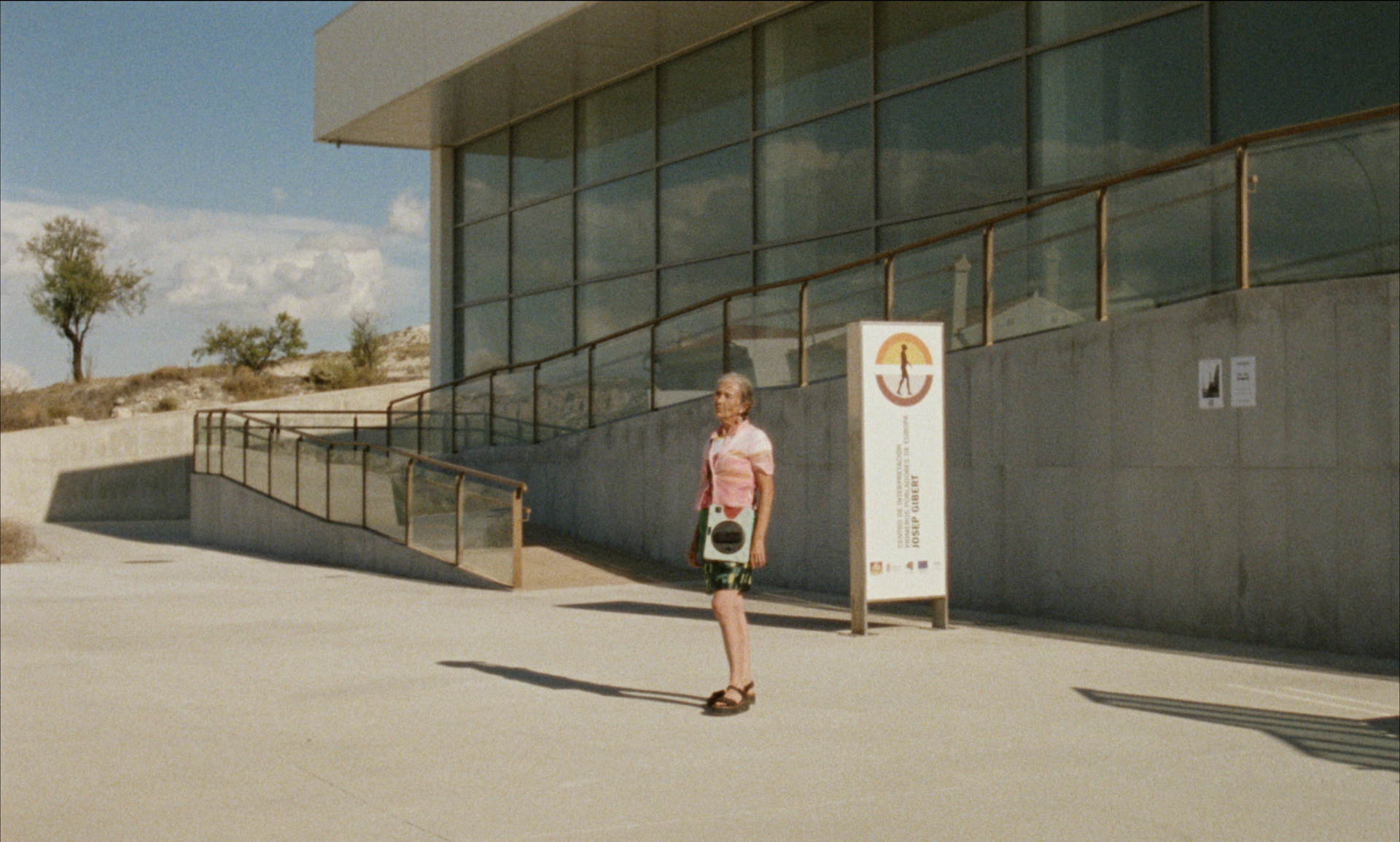 Una mujer está de pie frente a un edificio con ventanas de cristal, cerca de un letrero. Luz solar intensa.