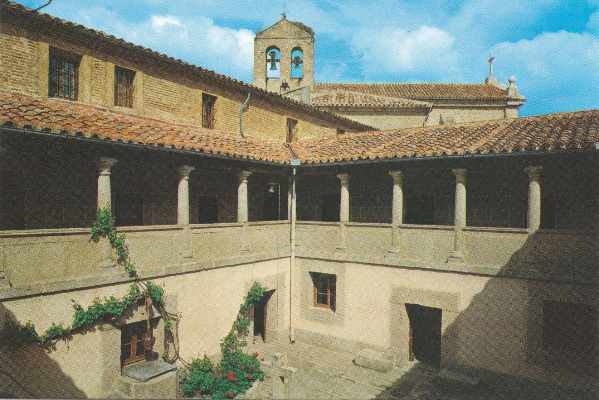 Patio con columnas de piedra, tejado de tejas rojas y campanario al fondo. Enredaderas verdes trepan por las paredes.