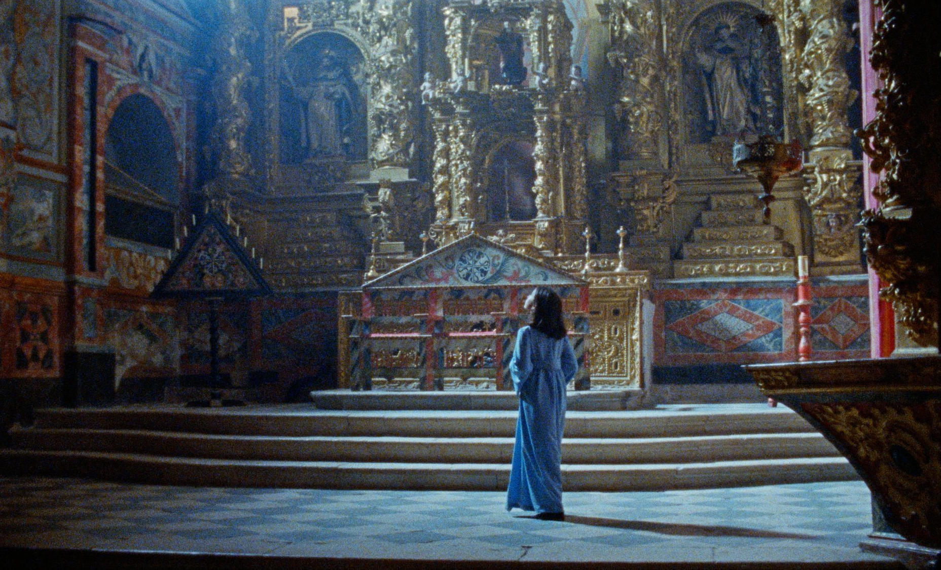 Una mujer vestida con un traje azul se encuentra de pie ante un altar dorado y ornamentado en una iglesia tenuemente