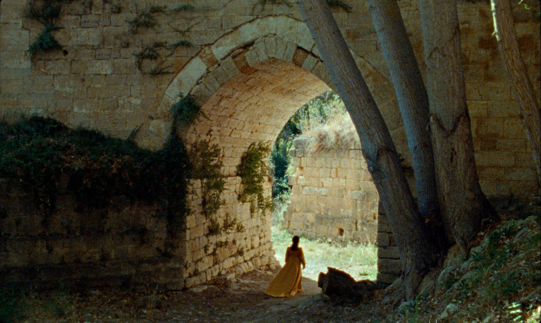 Una mujer vestida de amarillo camina a través de un arco de piedra; la luz del sol ilumina las paredes y la vegetación