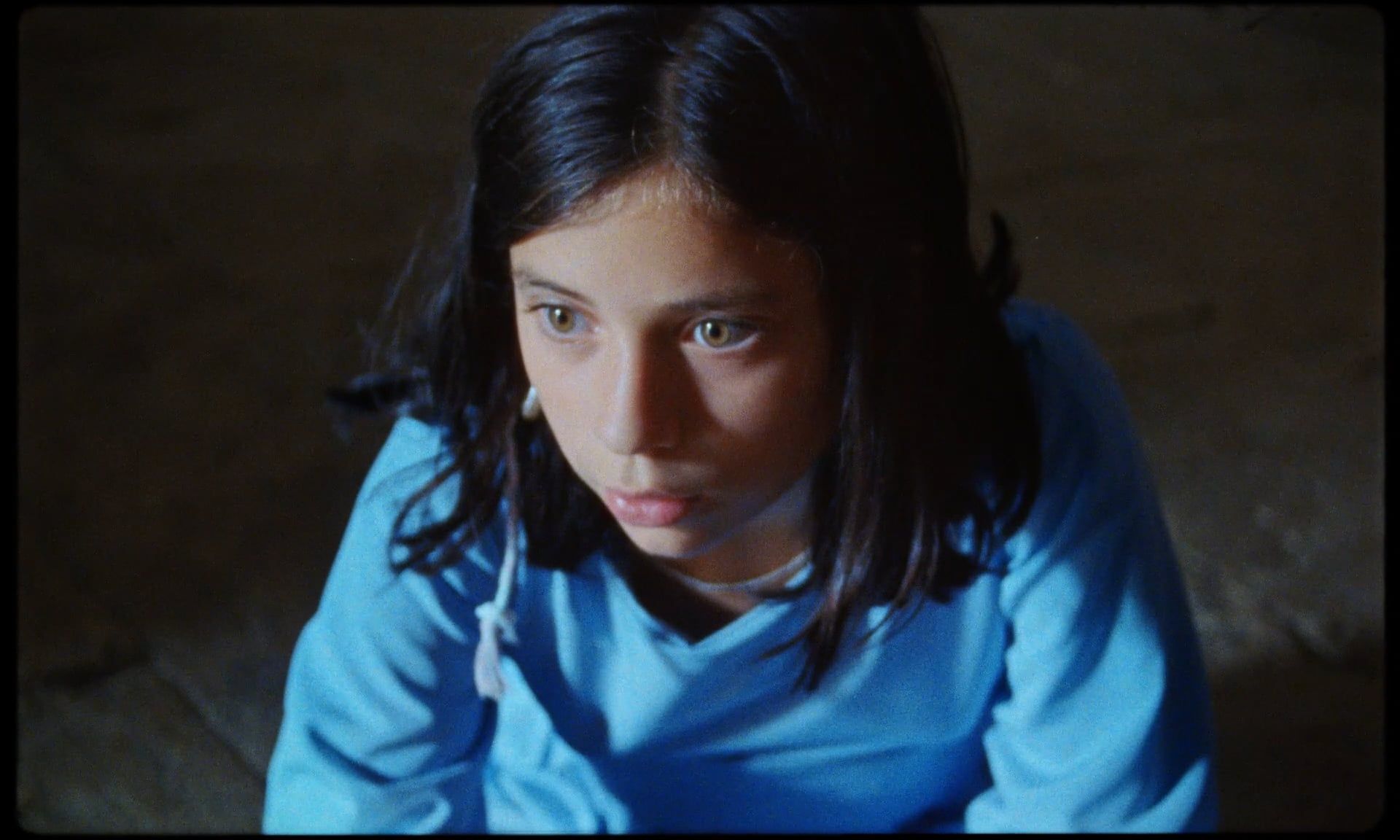 Una chica con camisa azul mira fijamente al suelo con expresión concentrada. Ambiente interior con luz tenue.