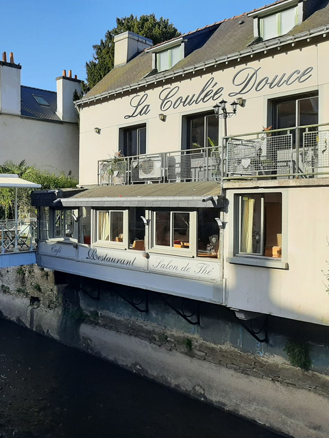 Un restaurant à la façade couleur crème nommé La Coulée Douce, avec un balcon donnant sur un canal par une journée ensoleillée.