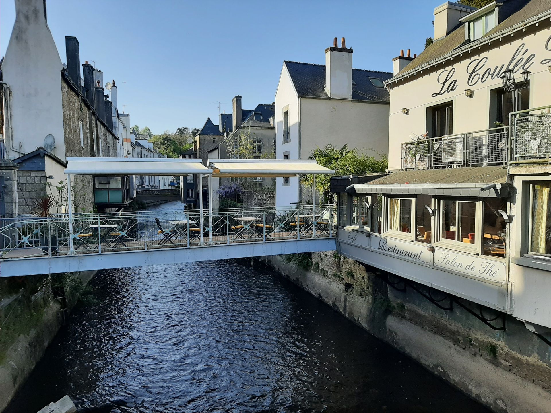 Une terrasse de restaurant construite au-dessus d'un canal, avec des tables, des auvents blancs et des bâtiments en pierre de part et d'autre.