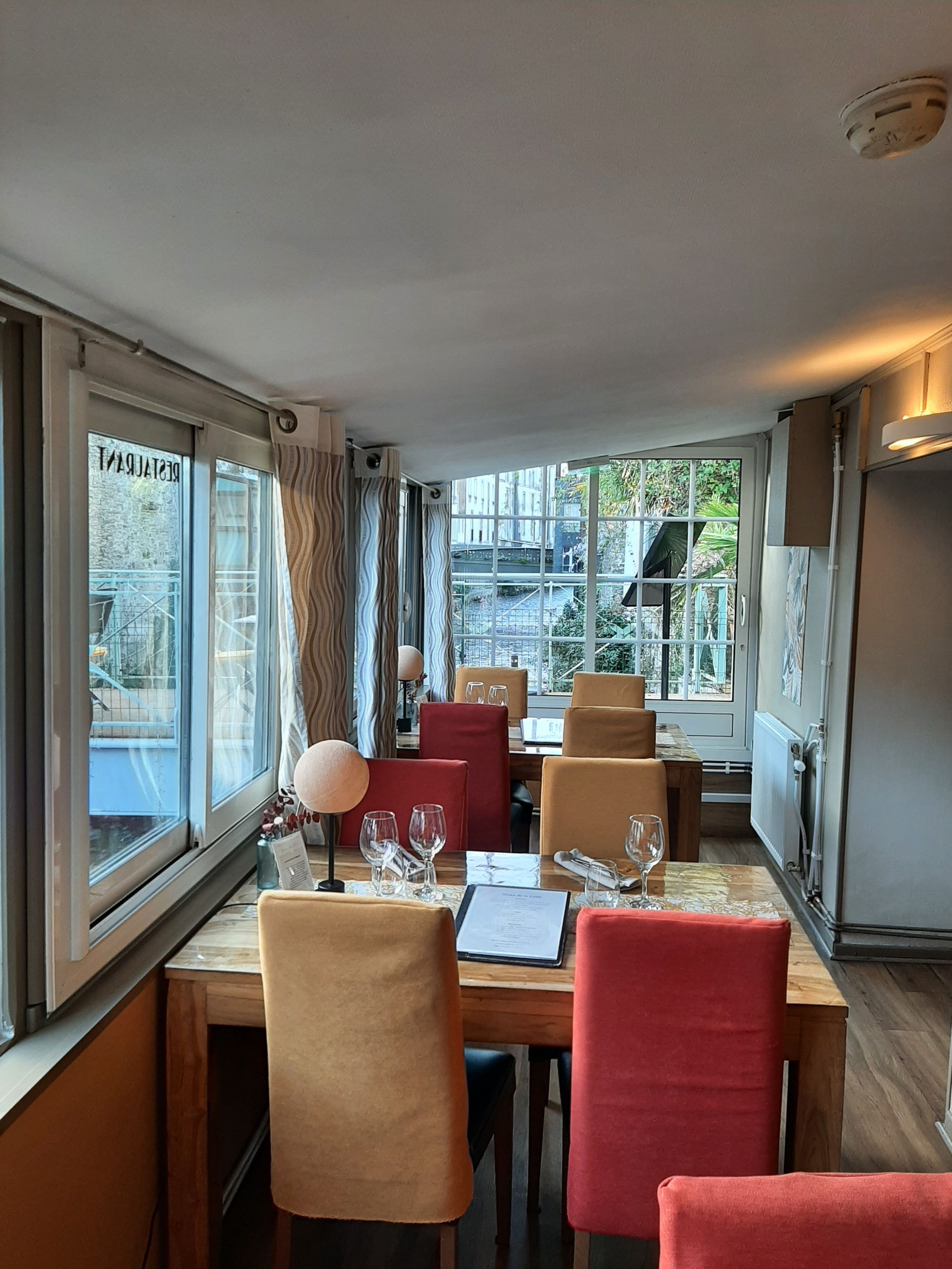 Coin repas avec tables et chaises en bois dans les tons de rouge et de beige, situé près de grandes fenêtres donnant sur les arbres.