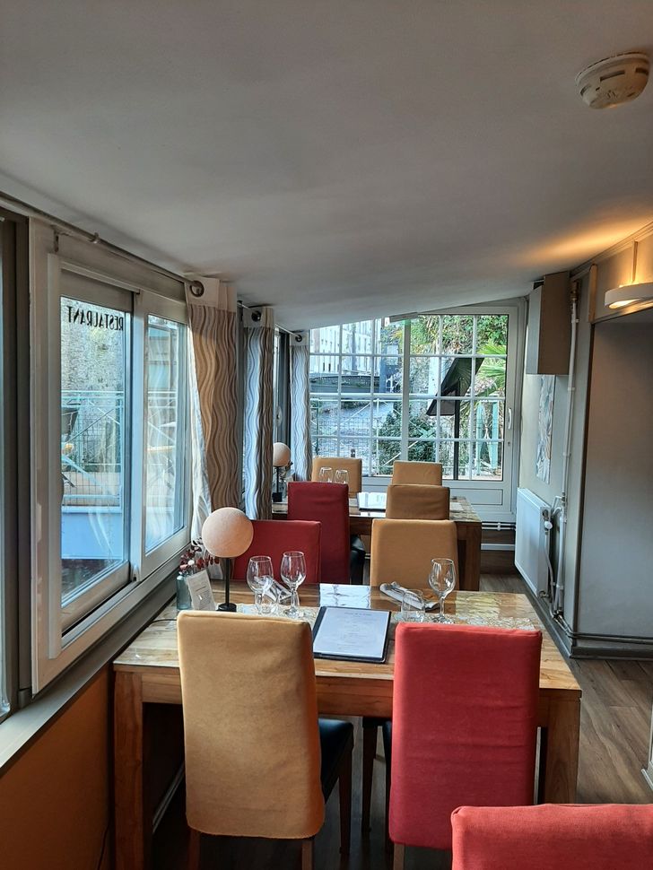 Coin repas avec tables et chaises en bois dans les tons de rouge et de beige, situé près de grandes fenêtres donnant sur les arbres.