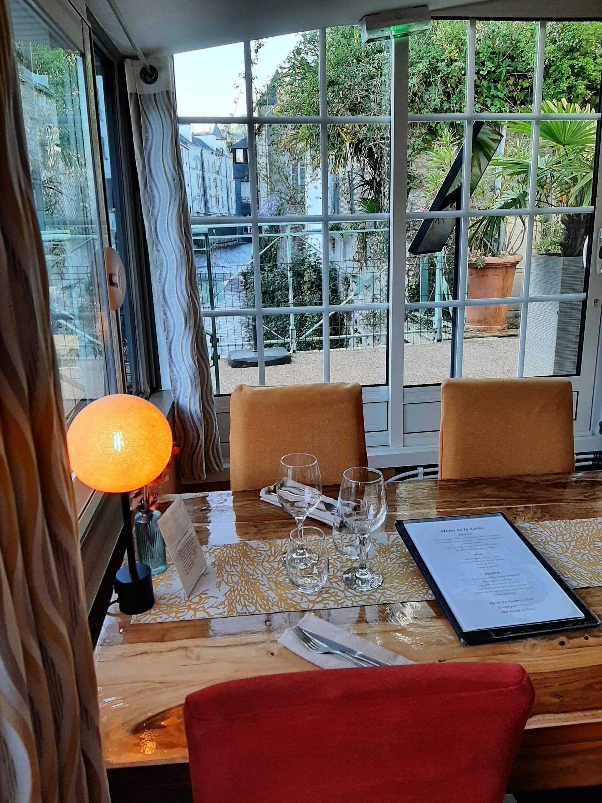 Une table dressée dans une véranda avec une lampe à la lumière chaude, des chaises, de la verrerie et un menu, donnant sur l'extérieur.