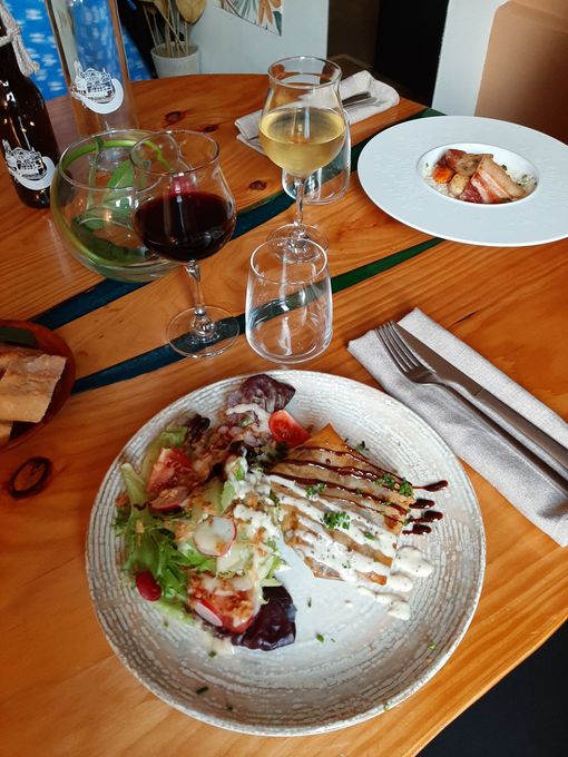 Une table à manger en bois dressée avec deux assiettes de nourriture, des verres de vin rouge et blanc, et des couverts sur une serviette blanche.