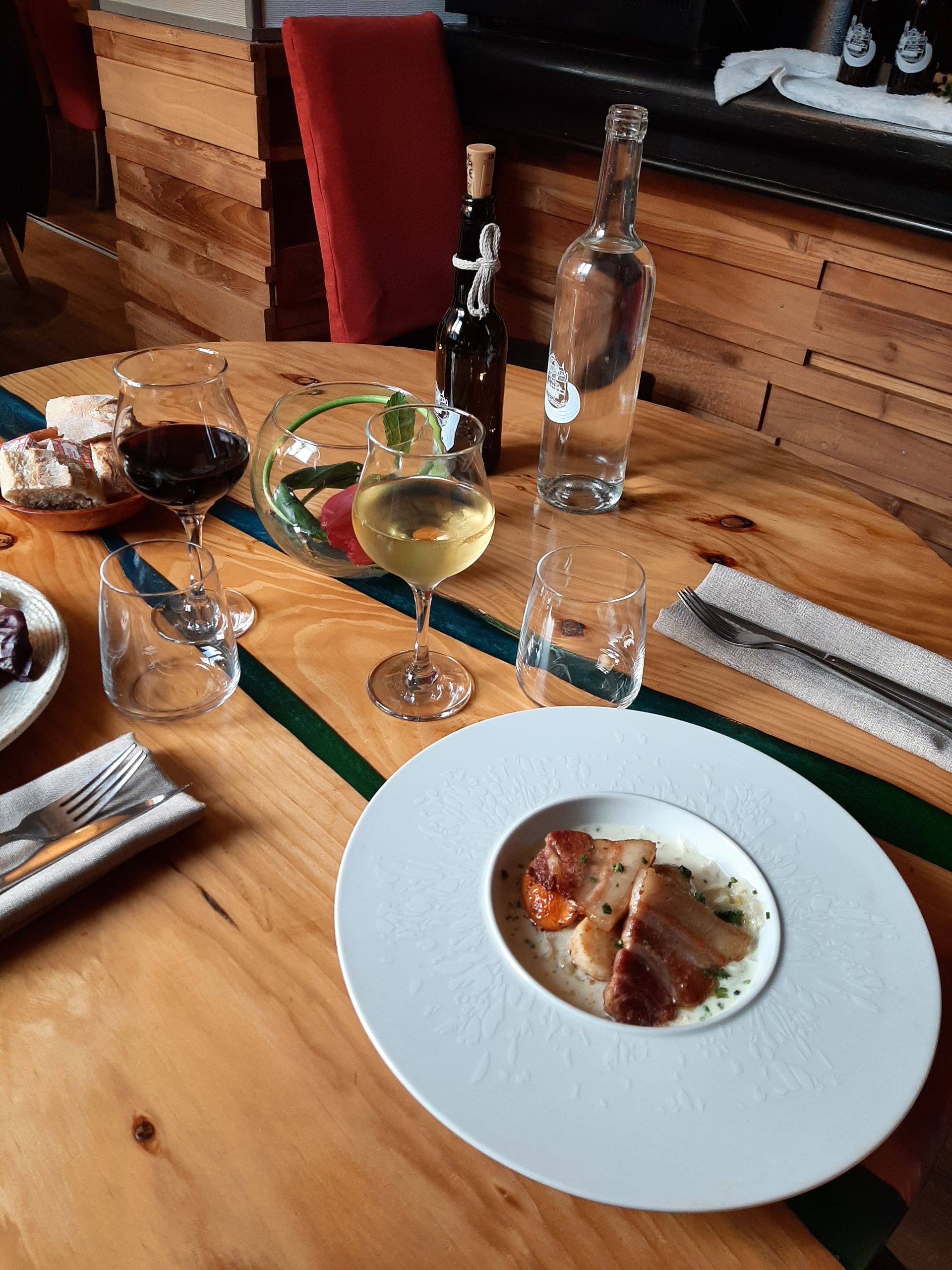 Une table en bois dressée pour un repas avec une assiette de porc tranché, des verres de vin, une bouteille d'eau et des petits pains.