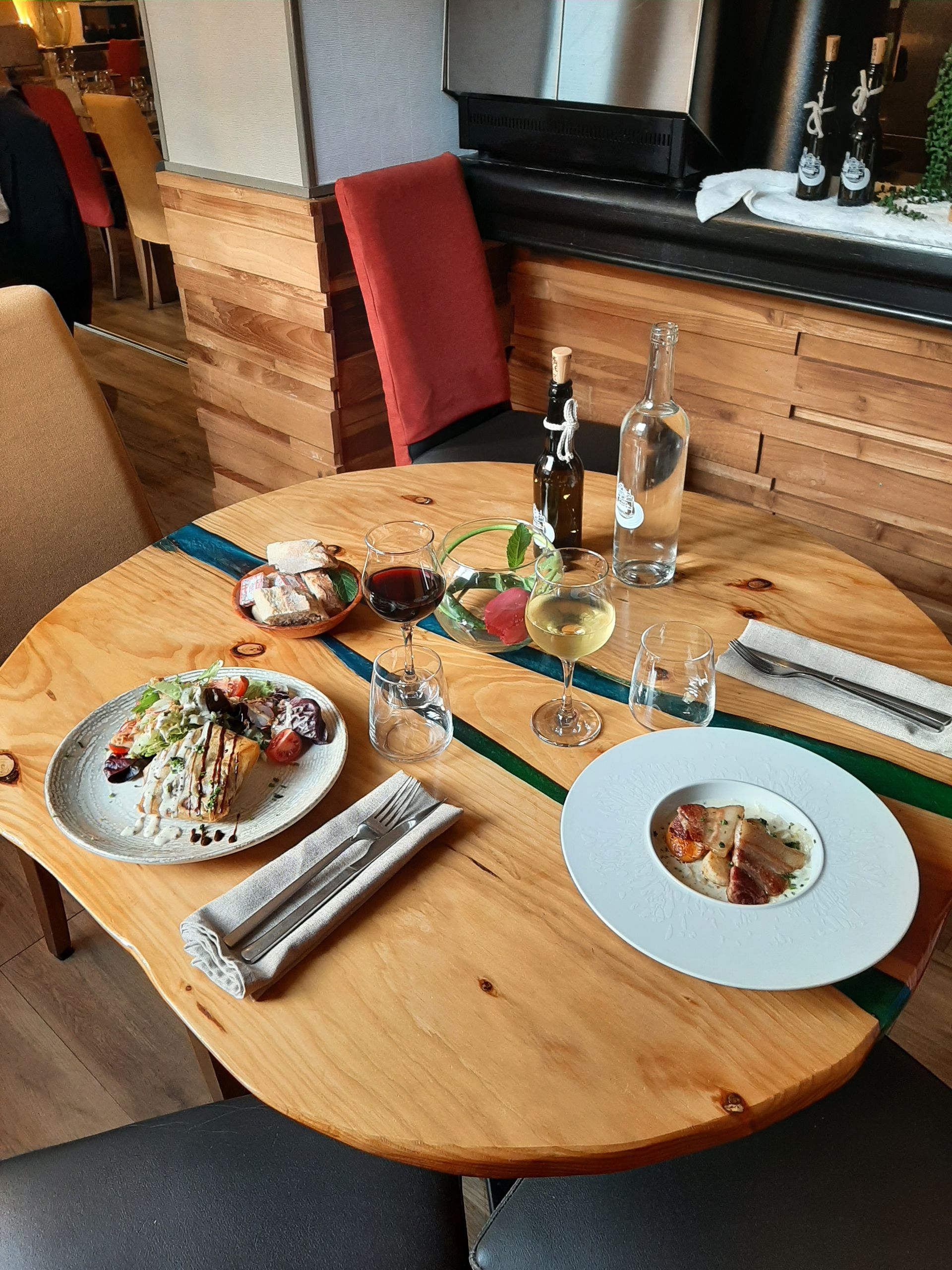 Dans un restaurant, une table ronde en bois est dressée pour deux personnes avec des assiettes de nourriture, des verres de vin, une carafe d'eau et des couverts.