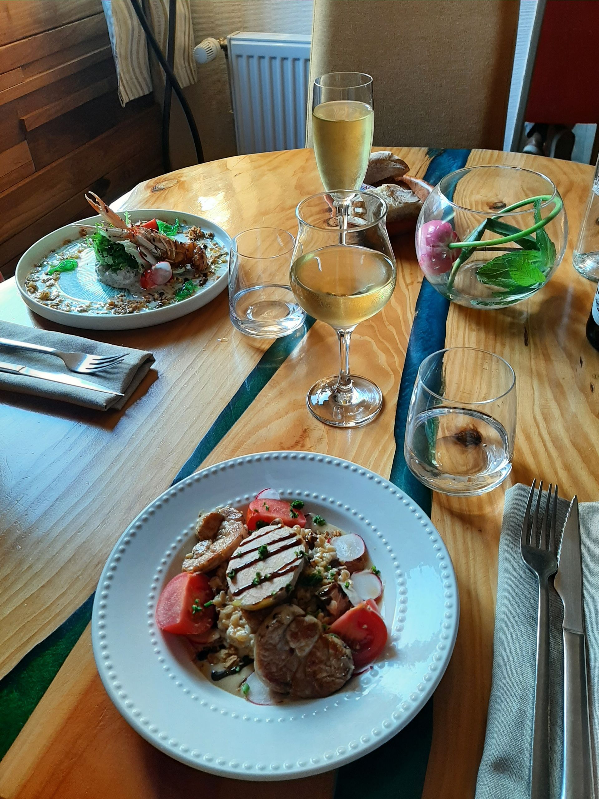 Une table dressée avec deux assiettes de nourriture, des verres de vin et un vase de fleurs, le tout sur une table en bois incrustée de résine bleue.