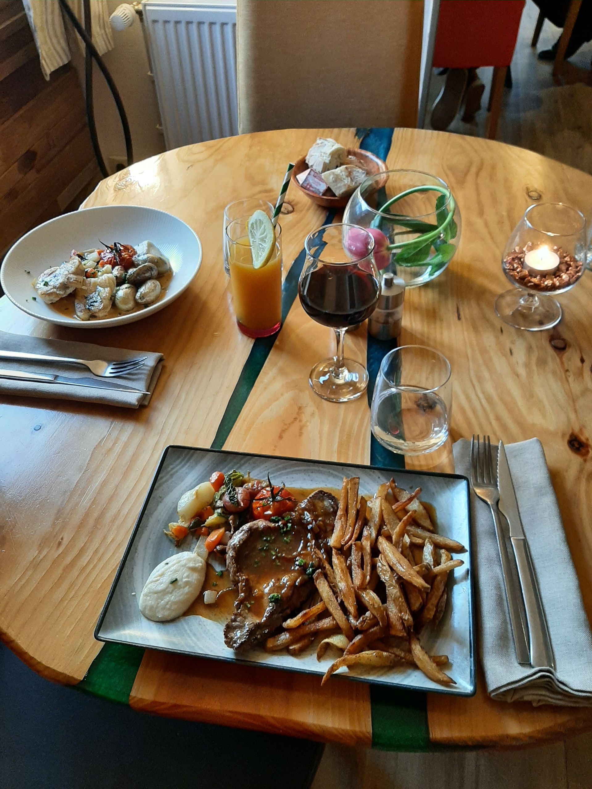 Une table en bois dressée pour deux, avec un steak-frites, un plat à base de pommes de terre, diverses boissons et une petite bougie allumée.