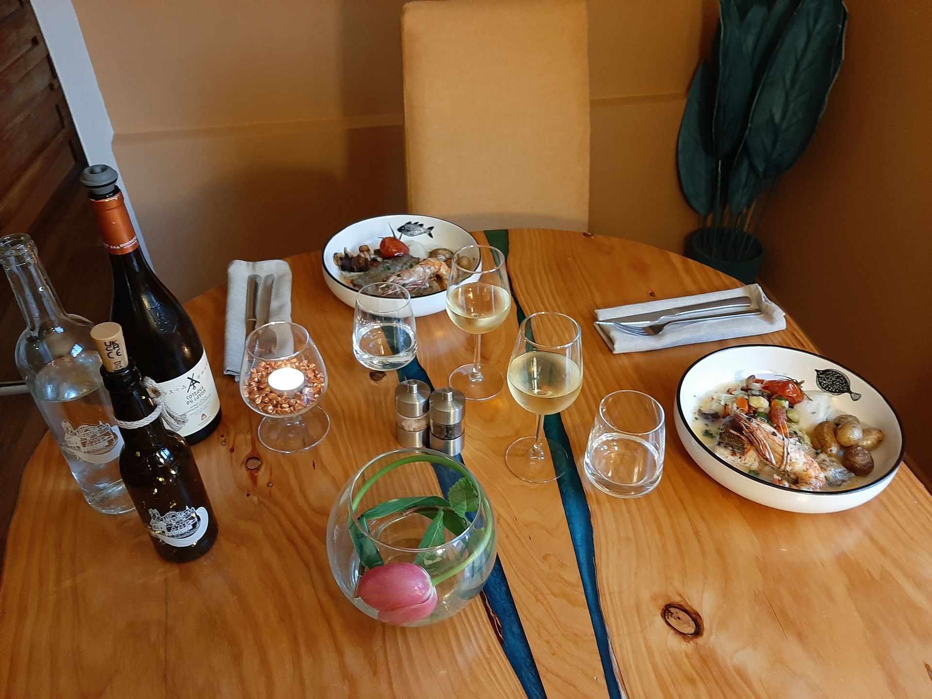 Une table en bois dressée pour deux, avec des assiettes de nourriture, du vin, de l'eau, des couverts et un petit vase à fleurs.