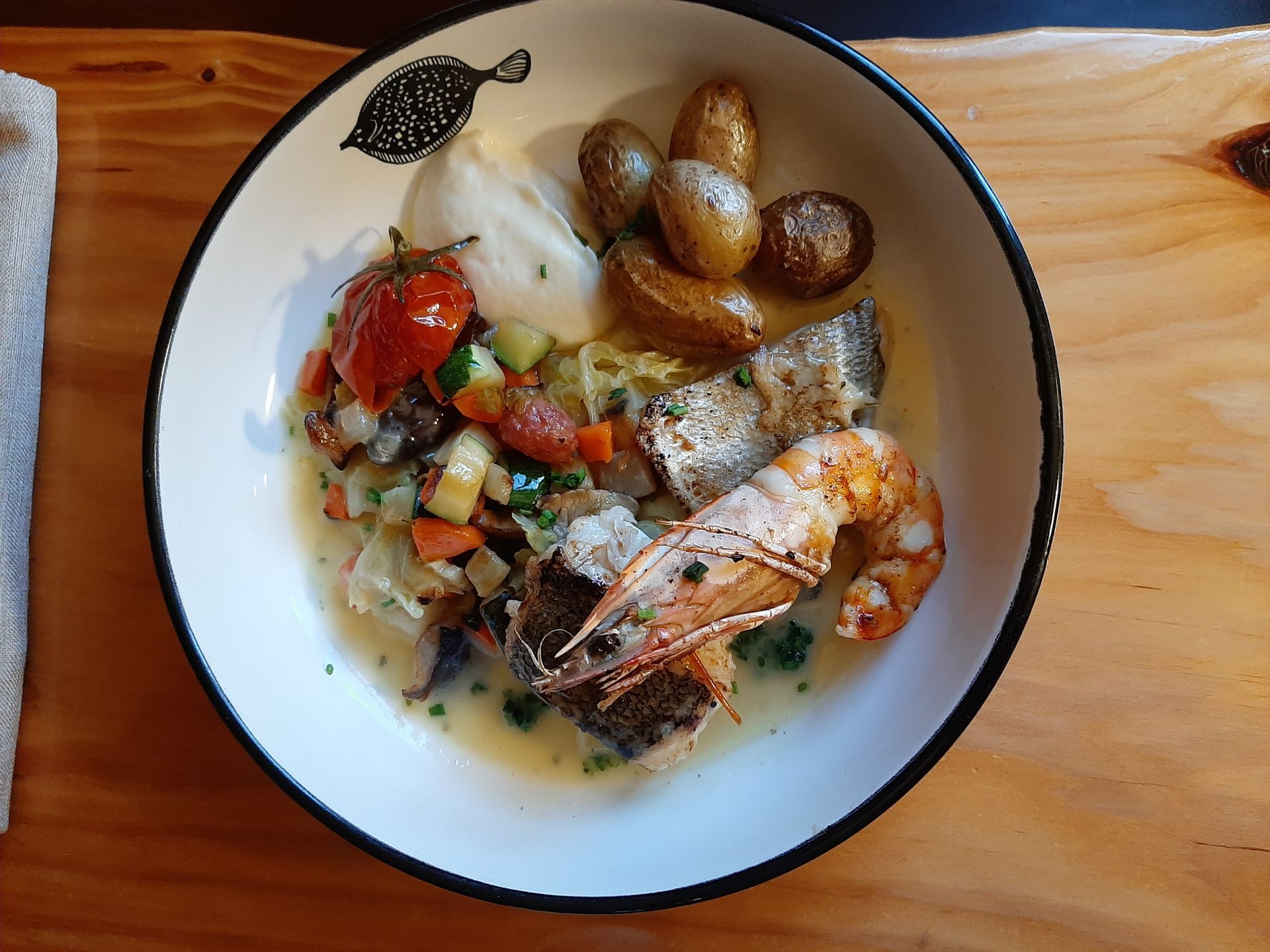 Un plat composé de poisson, d'une grosse crevette, de pommes de terre rôties et d'un assortiment de légumes dans une sauce crémeuse, le tout servi sur une planche en bois.