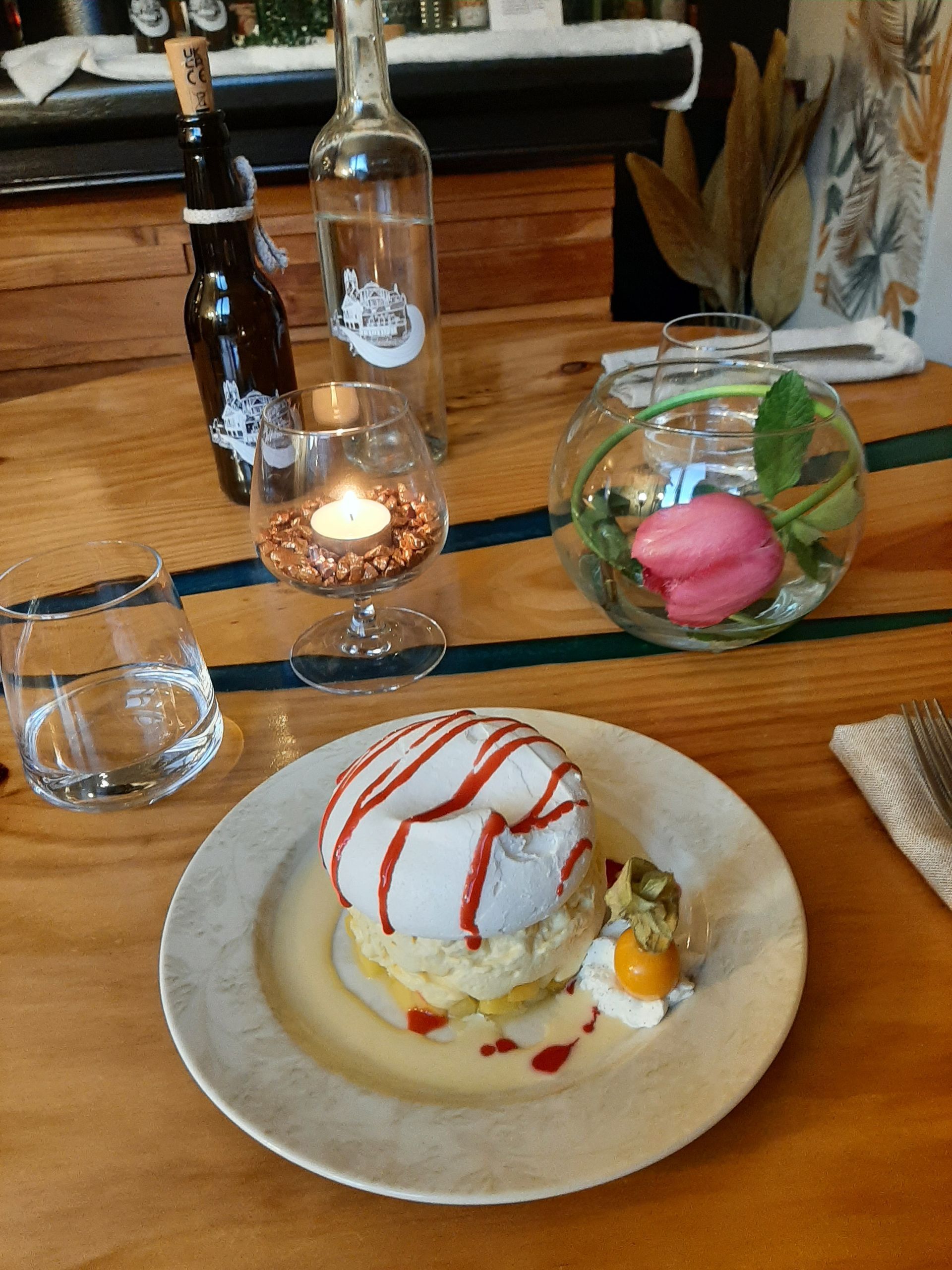 Un dessert à l'assiette, composé de meringue blanche et de sauce rouge, accompagné d'un verre d'eau, d'une bougie allumée et d'une fleur dans un bol.